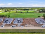 Row of white houses under construction along a lush golf course with greens and sand traps beyond the row of homes, under a clear sky, aerial view.