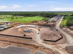 Construction site with dirt roads, equipment, and building foundations next to a green golf course complex and parking lot.