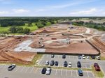 Construction site with dirt roads, heavy equipment, and soil piles, adjacent to a parking lot and green fields under a blue sky.