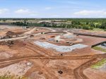 Aerial view of a large construction site with dirt, equipment, and a concrete foundation forming at the center, with a parking lot on the right and greenery in the background.
