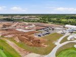Aerial view of a construction site with heavy machinery, piles of dirt, and a parking lot beside a green landscape and golf course fairways in the foreground.