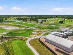 Aerial view of a expansive golf course with green fairways, sand traps, and water features, bordered by trees and a large white facility in the foreground