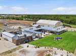 Aerial view of an event venue under construction: large white tent, metal buildings, paved roads, and a manicured lawn.