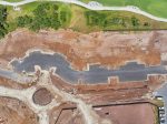 Aerial view of a construction site with a curved paved road under construction, brown dirt, and a green grassy area with trees at the top; a circular turn area at the bottom right.