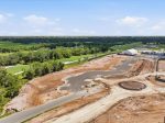 Aerial view of a construction site beside a golf course and green fields, with a large white tent building and cleared earth nearby.