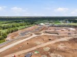 Aerial view of a construction site with dirt roads, earth piles, and a large white temporary building next to a green golf course and ponds in the distance.
