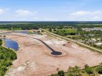 Aerial view of a curved road under construction on brown soil, with small ponds and surrounding green fields.