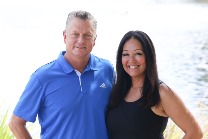 A smiling couple standing side by side outdoors by a lake, wearing casual clothes.